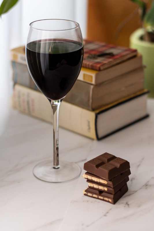 Ein Glas Wein und Schokolade, typische Trigger für Migränepatienten, stehen vor einem Stapel Bücher.
