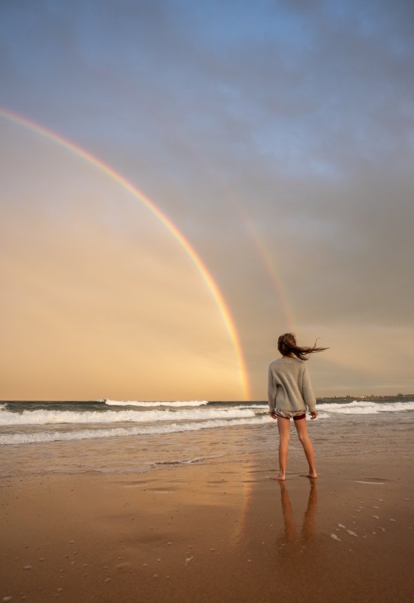 Frau am Strand mit Regenbogen. Zu Beginn einer Migräneattacke kann ein entspannender Spaziergang eine Attacke abwenden.