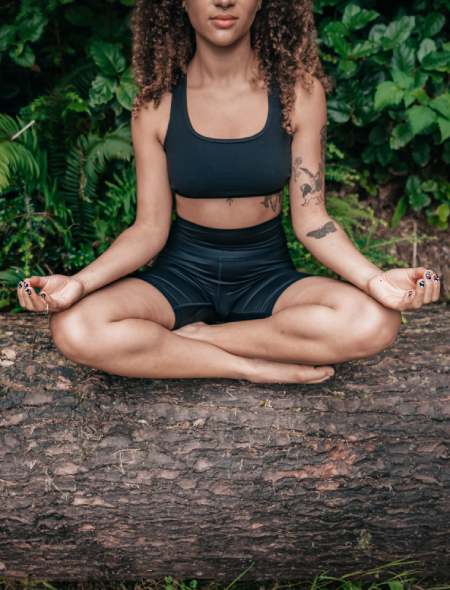 Frau in schwarzer Sportkleidung meditiert draußen in der Natur auf einem Baumstamm sitzend. Meditation hilft zur Vorbeugung gegen Migräne